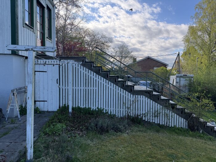 Sliten utomhustrappa i betong vid trähus med rött dörrparti, omgiven av vit staket och grönska, en del av äldre byggprojekt.