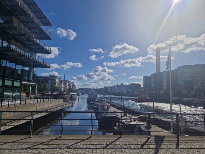 Stadskanal med båtar, moderna byggnader på sidorna och molnig himmel med solstrålar.