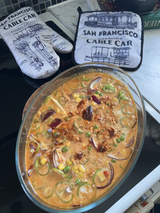 Ugnsform med lax i kokosmjölksås med rödlök, ärtor, majs, paprika och kryddor på en spis. Grytlappar med San Francisco Cable Car-tema.