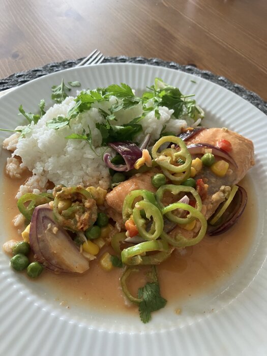 Tallrik med ugnsbakad lax i kokosmjölk, garnerad med rödlök, ärtor, majs, paprika, och serverad med ris och koriander.