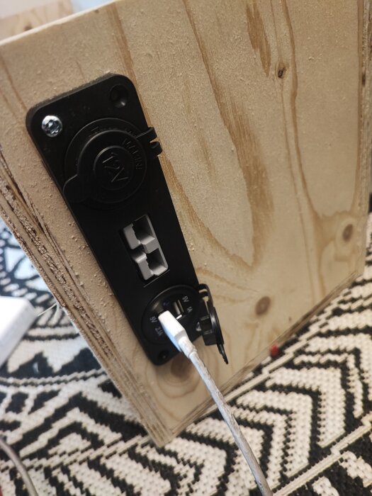 En träpanel med ett monterat 12V-uttag och en ansluten vit kabel på en mönstrad matta.