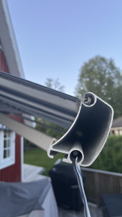 Profilbild av en öppen aluminiumprofil på en husfasad, potentiellt del av en markis eller liknande konstruktion, med trädgård i bakgrunden.
