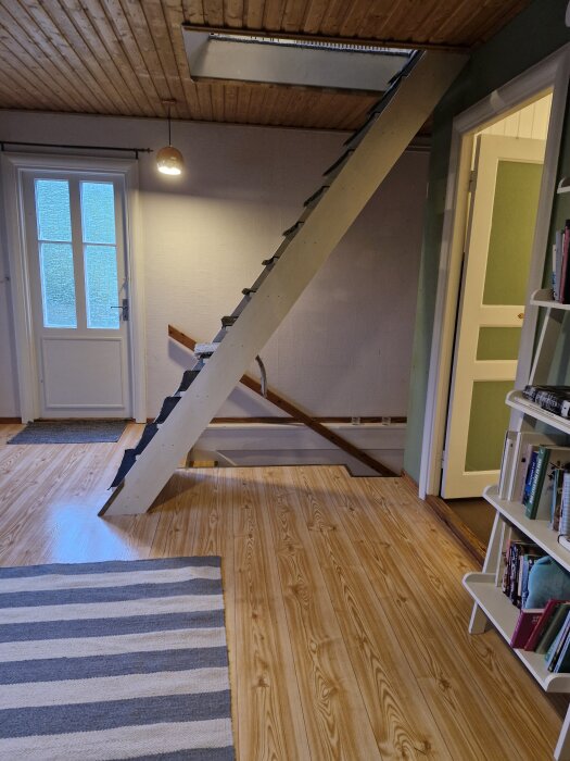Stegtrappa i ett äldre hus, placerad framför dörren till badrummet. Inga räcken syns, vilket ger en osäker känsla. Träpanel i tak och golv.