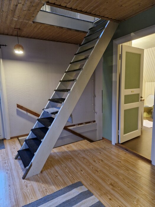 Brant vindstrappa i ett äldre hus, placerad intill en öppen dörr till badrummet, med träväggar och tak samt trägolv.