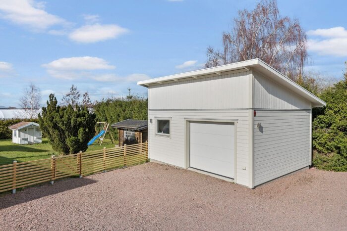 Vitt garage med port på långsidan, omgiven av staket och trädgård med lekstuga och gungställning i bakgrunden.