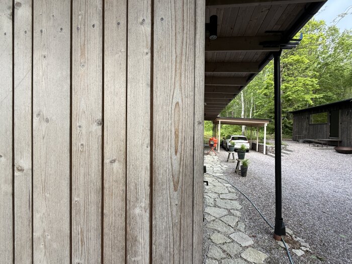 Träfasad med behandlade plankor intill en stengång, under ett tak, med en bil parkerad i bakgrunden nära grönska.