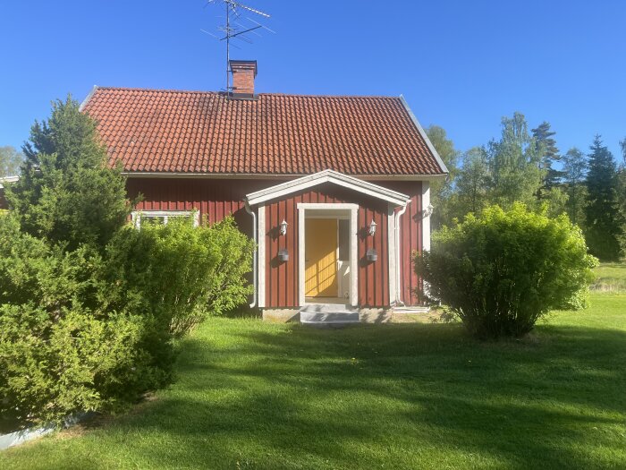 Rött trähus med rött tegeltak, gröna buskar och gräsmatta i solsken.