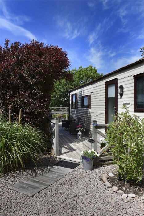 En villavagn i Kungshamn med stor terrass av tryckt virke, omgiven av grönska och blommor under en klarblå himmel.