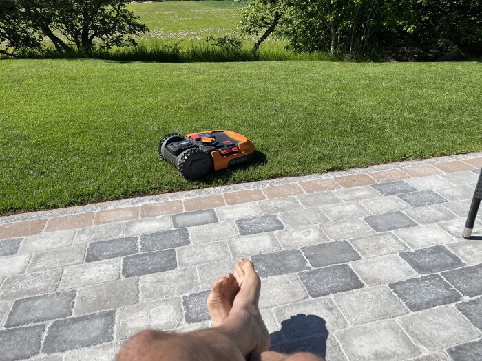 Robogräsklippare på en välklippt gräsmatta nära en stenlagd uteplats med en persons fötter synliga i förgrunden.