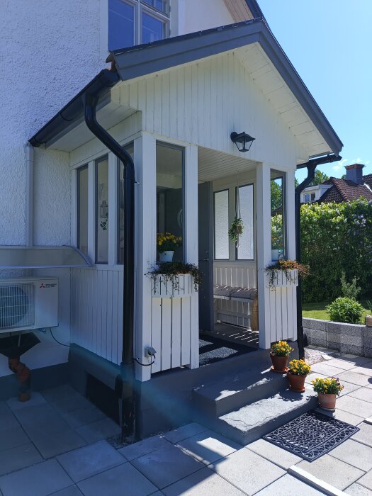 Vit entré med plantering vid ytterdörr, betongtrappa med blommor i krukor och asfalt utanför. Huset har vita paneler och svarta stuprör.