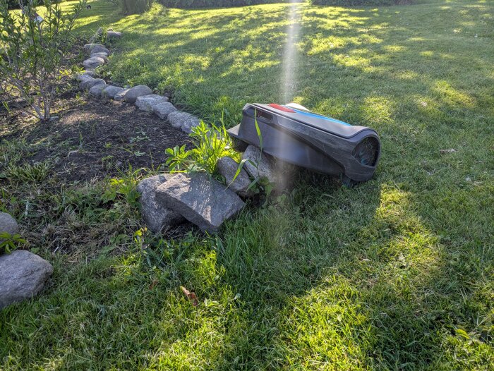 Robotgräsklippare fastkörd på en sten i en trädgård, omgiven av gräsmatta och kantstenar.