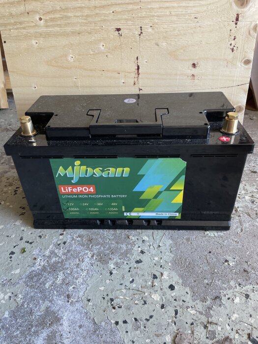 Litiumjärnfosfatbatteri 12V 100Ah med etikett "Mjdsan" står på betonggolv framför trävägg.