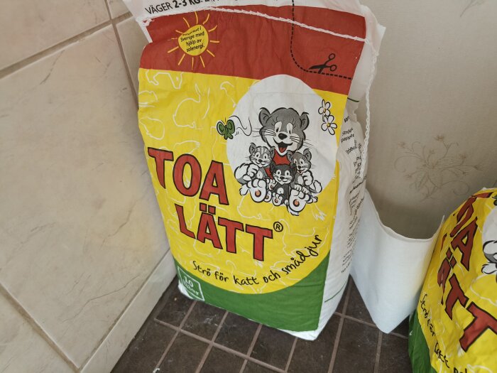 Ett paket av kattströ "Toa-Lätt" står på ett badrumsgolv. Förpackningen har en tecknad bild av en mus med ungar.