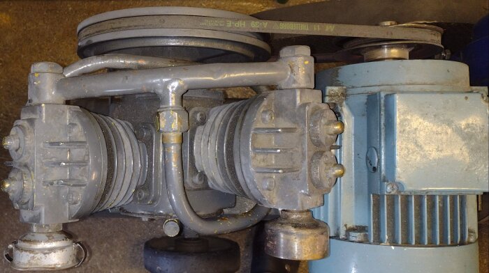 Gammal kompressor med remdrift, visar två cylindrar och en koppling till elektrisk motor, frågeställning om vevaxeltätningstyp och byte.