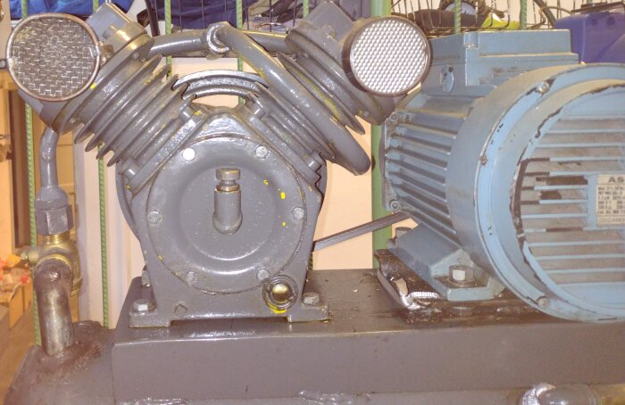 Grå kompressor med två cylindrar och kraftig motor, eventuell vevaxeltätning synlig.