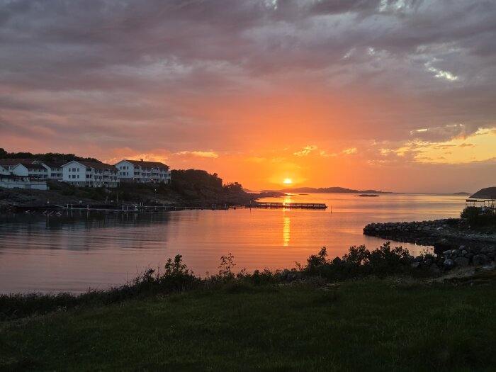 Solnedgång över havet med flera byggnader på en klippa till vänster och en brygga som sträcker sig ut i vattnet, himlen är färgad i orange och rosa.