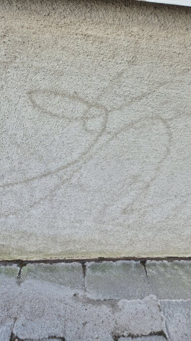 Ljusspräcklig putsvägg med ojämna mörkgrå linjer som ser ut som klotter, orsakade av högtryckstvätt, mot stenbelagd mark.