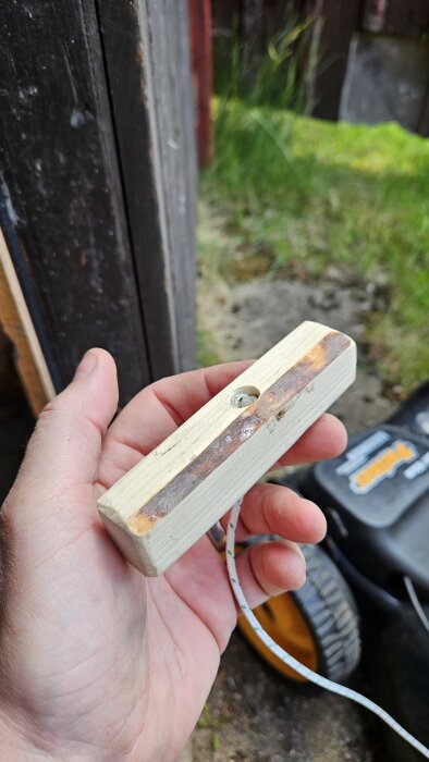 Hand håller i träbit med fäst startsnöre framför gräsklippare och grönska i bakgrunden.