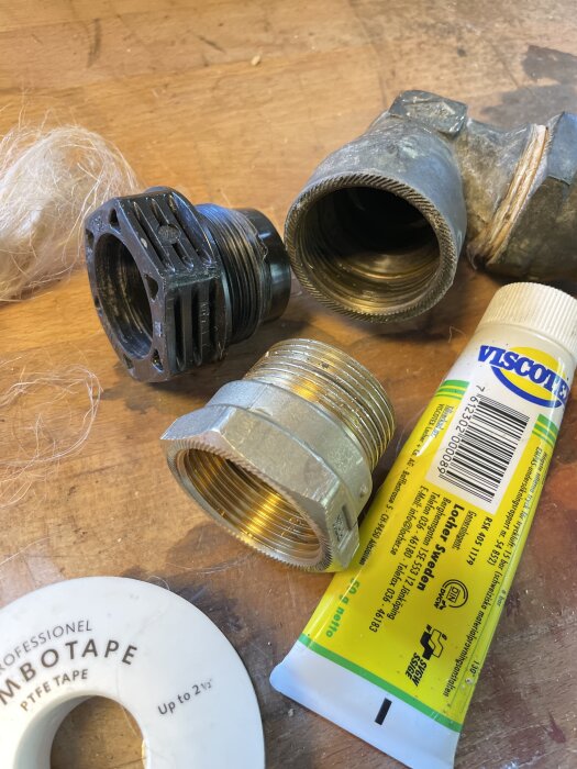 Vattenledningstillbehör med vinkelkoppling i mässing, en svart plastdel, lin, pasta och PTFE-tejp på ett arbetsbord.