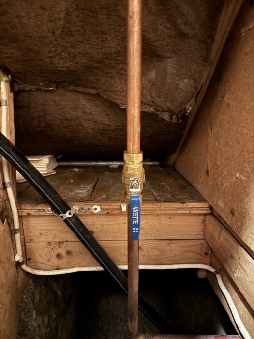 Vattenledning i snickrat träutrymme med blå etikett "VATTEN", kopplad med kopparrör och ventiler, installeras under trångt utrymme.