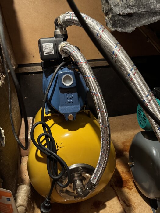 Gul hydroforpump med blå anslutning och två metallomspunna slangar. Pumpen är placerad i ett trångt utrymme.