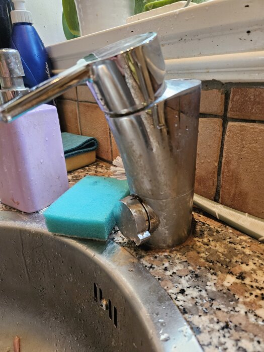 Köksblandare med avbruten pip monterad på en diskbänk, med en blå svamp och rengöringsartiklar i bakgrunden.