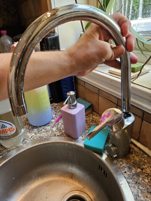 En hand håller en avbruten pip från en köksblandare ovanför en diskbänk med rengöringsprodukter och en svamp.