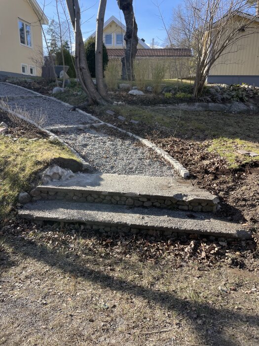 Utomhustrappa i två steg med ojämn yta och sprickor, omgiven av grusgång och trädgårdsmiljö.