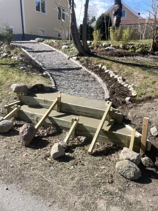 Utomhustrappa under konstruktion med synliga brädstöd och omgivande stenar, belägen intill en gångstig i en trädgård.