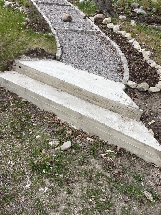 Utomhustrappa i två steg med ojämn yta, färgskiftningar och små sprickor, omgiven av grus och stenar.