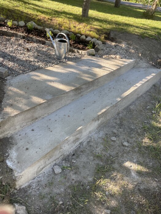 Betongtrappa med två steg och ojämn yta samt färgskiftningar, omgiven av grus och gräsmatta. En vattenkanna står på gruset bredvid trappan.