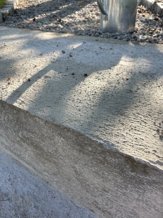 En uteplats med ojämn betongyta och färgskiftningar. Runt den syns grus och skuggan av en vattenkanna i metall.