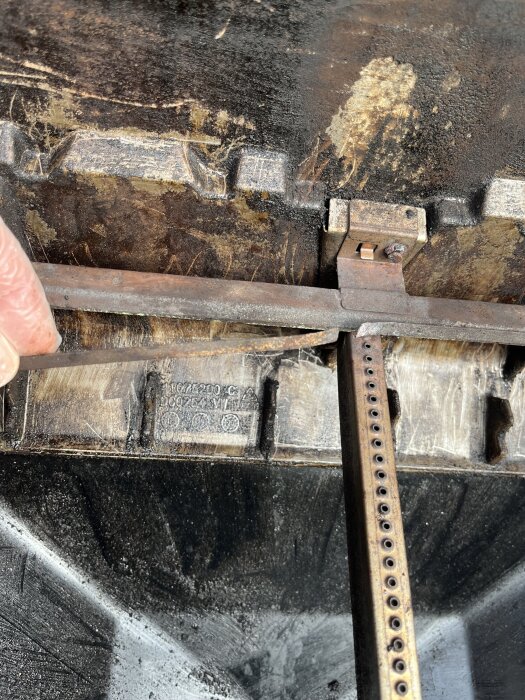 Närbild av insidan på en grill, visar rostiga brännarrör och en lossnad list, med bränd och smutsig yta.