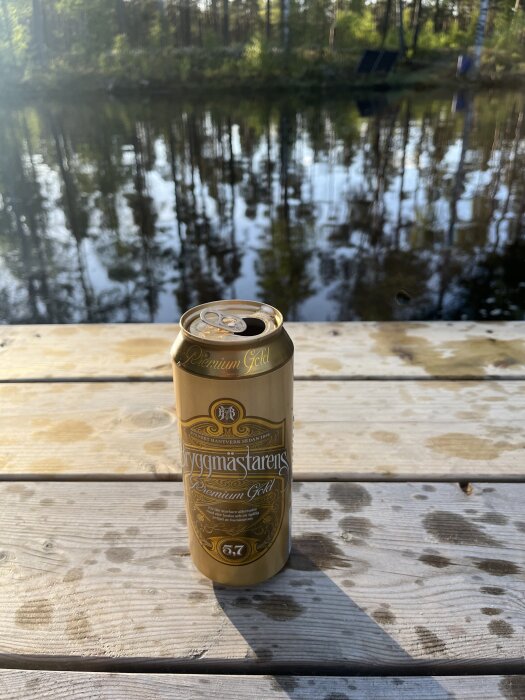 En ölburk med bryggmästarens premium gold på en brygga vid en spegelblank sjö och träd i bakgrunden.