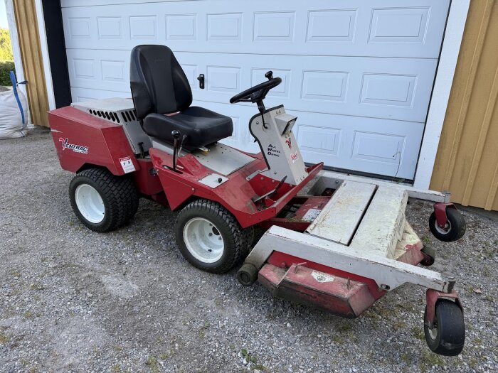 Röd åkgräsklippare av märket Ventrac med nytt säte, parkerad framför en garageport.