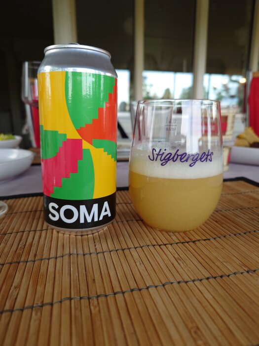 En ölburk med färgglad design bredvid ett glas ölfyllt glas med skum på bambuunderlägg.