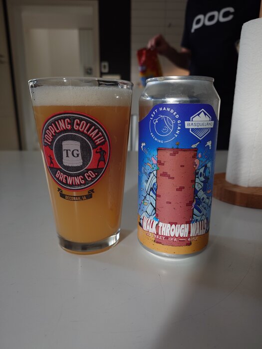 Ett glas öl med logotypen "Toppling Goliath Brewing Co." och en burk öl med etiketten "Walk Through Walls" på ett bord.