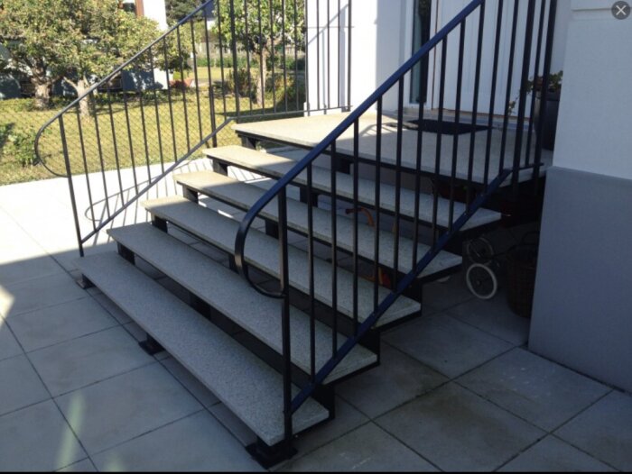 Utomhustrappa med granitsteg och svart räcke, leder upp till entré med stenbeläggning runt, soligt väder och grönska i bakgrunden.