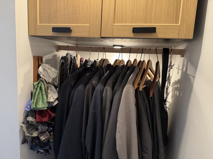 Klädhängare med kavajer och skjortor, träskåp ovanför, samt hylla med vikta kläder och accessoarer i en walk-in-closet.