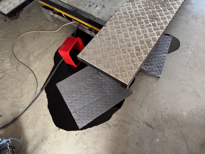 En verkstadsgolvyta med metallplattor, oljefläck och absorberande mattor samt en röd tratt.