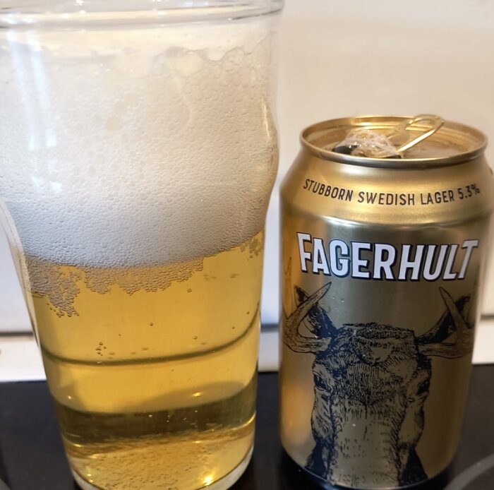 Glas med skummande lageröl bredvid en burk Fagerhult Stubborn Swedish Lager, 5,3% alkohol, på en bänk.