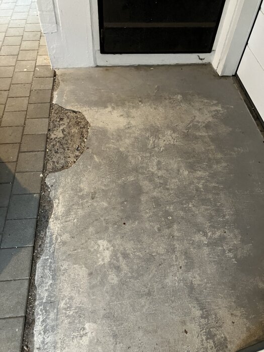 Skadad cementyta vid garageinfart, flisor saknas längs kanten, angränsar till stenplattor.