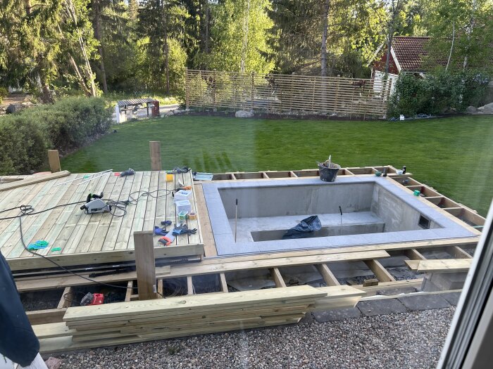 Byggprojekt med ofärdig pool och ny trall på gården, byggmaterial och verktyg utspridda, omgiven av grönt gräs och träd.