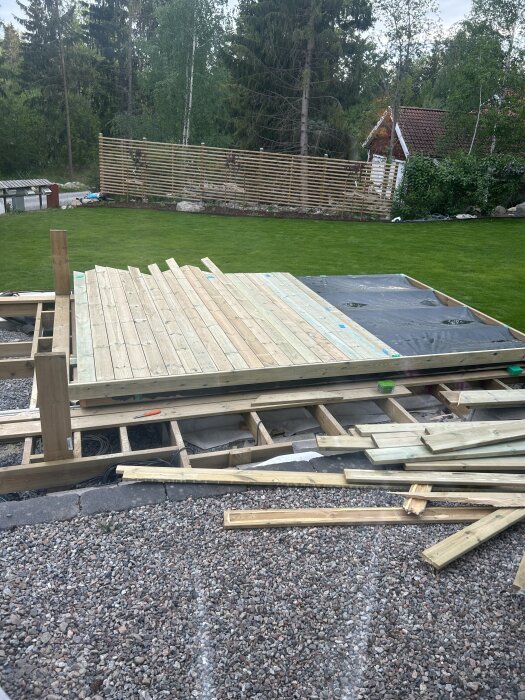 Pooltak under byggnation i trädgård, med trall på ramverk och skyddande EPDM-dukar i mitten. Lösa trallbrädor och verktyg på marken.