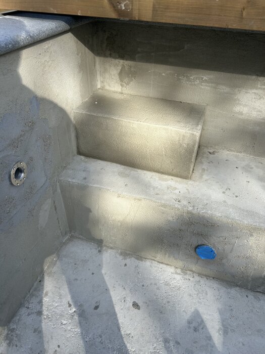 Betongtrappsteg i en under konstruktion av en pool. Två steg, den nedre med pluggad öppning. Skugga från två personer syns på betongytan.