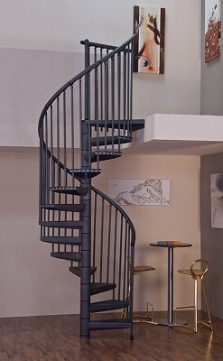 En svart spiraltrappa i ett modernt inrett rum med konst på väggarna, stående på ett brunt golv bredvid ett litet bord och stolar.