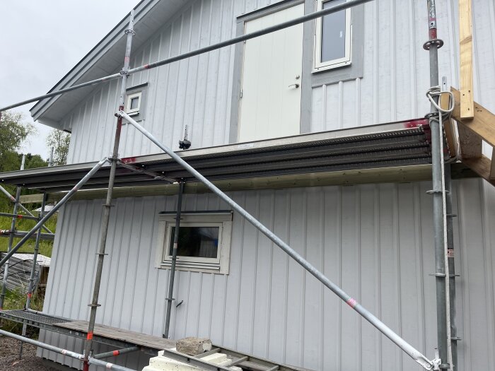 Byggställning och fastighet med monterad dörr och fönster, regel för framtida altan synlig på övervåningen.