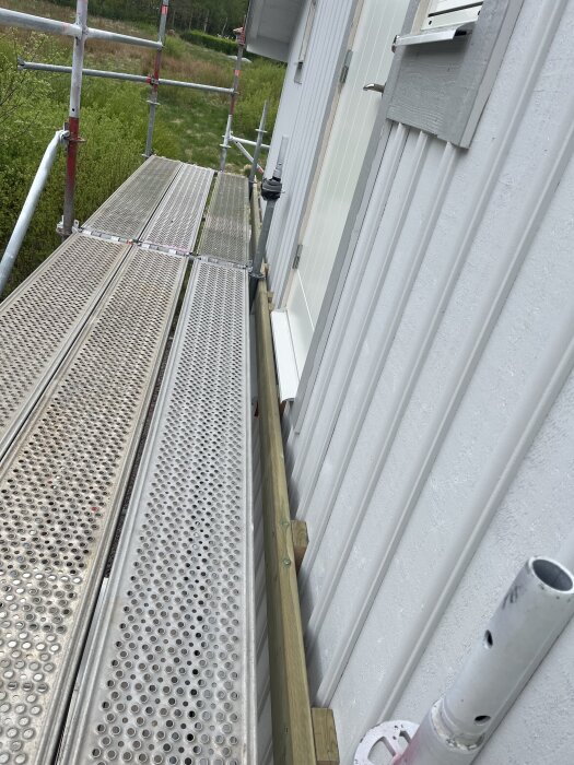 Byggställning med plattformar vid en vit husvägg och en träregel för framtida altankonstruktion.