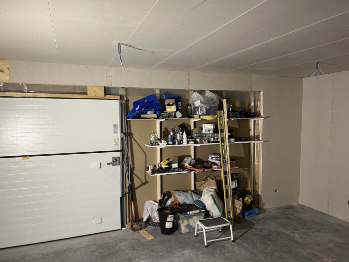 Gipsad vägg med monterad central, hylla fylld med verktyg och material samt en stege i ett garage eller arbetsrum.