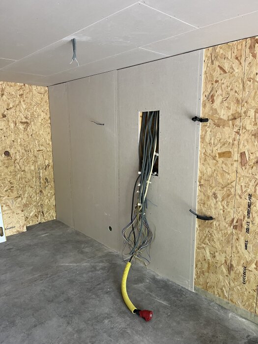 Byggvägg med gips och träskivor, exponerade elektriska kablar för montage av centralen; en gul slang med röd ände ligger på grå betonggolv.